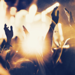 Rear view of large group of unrecognizable people at concert. Their hands are in the air, clapping. Beige stage lights in the background. Two people in foreground are released.